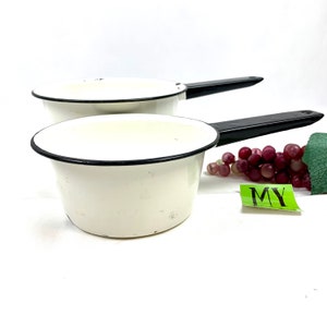 Può includere: Due pentole di smalto bianco con manici neri. Le pentole sono su una superficie bianca con un grappolo di uva rossa e una foglia verde sullo sfondo. La parola "MY" è scritta su un adesivo verde.