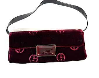 Puede incluir: Un bolso de mano de terciopelo burdeos con una correa negra. El bolso presenta un patrón repetido de un logotipo estilizado y un cierre rectangular con un acento burdeos. El bolso es de estilo clutch.