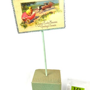 May include: An Easter-themed decorative sign featuring a vintage illustration of a chick in a red carriage pulled by rabbits. The sign has a glittery border and reads "Joyous Easter Memories with Greetings Sincere."