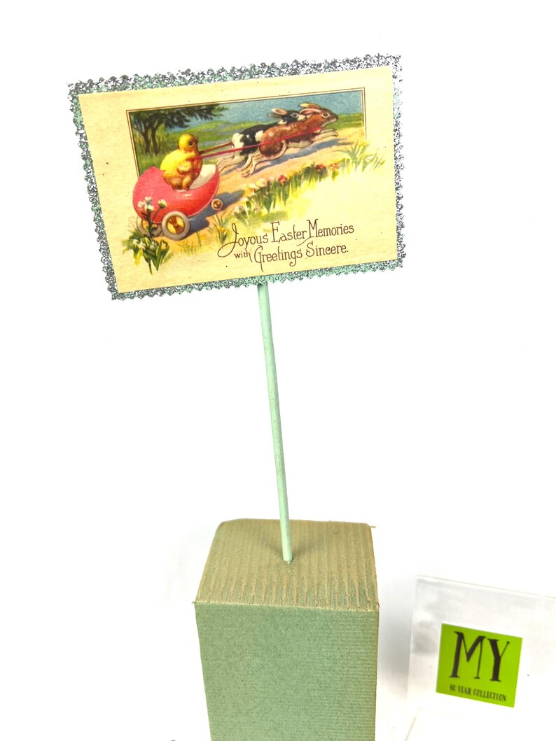 May include: An Easter-themed decorative sign featuring a vintage illustration of a chick in a red carriage pulled by rabbits. The sign has a glittery border and reads "Joyous Easter Memories with Greetings Sincere."
