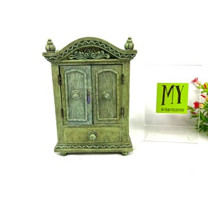 May include: A small, green wooden cabinet with two doors and a drawer. The cabinet has a decorative top and ornate details. The cabinet is a vintage style and has a distressed finish.