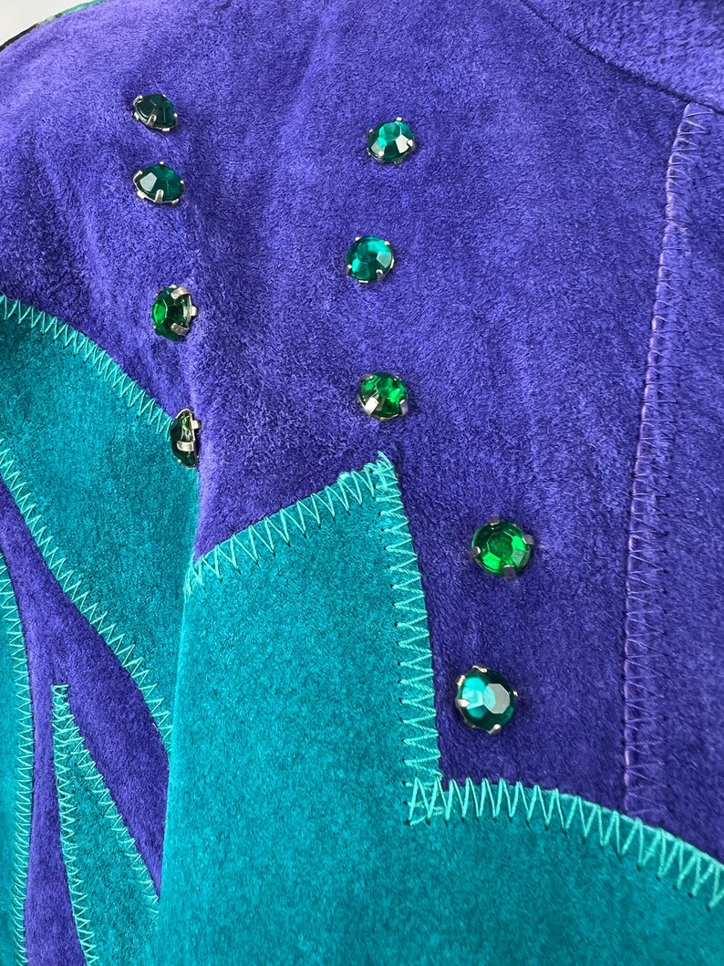 May include: A purple and teal suede jacket with green rhinestone embellishments. The jacket has a geometric design with a zig-zag stitch detail.