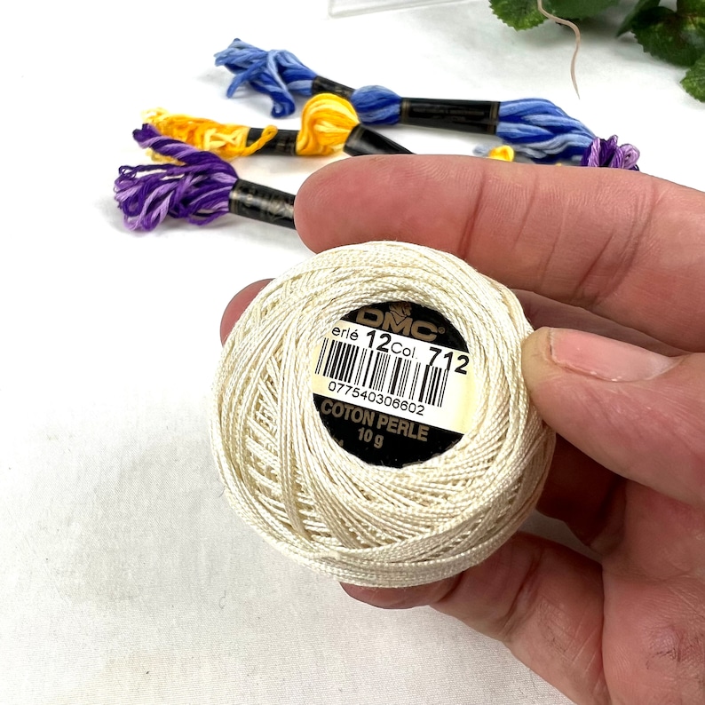 May include: A white DMC embroidery floss spool with a label that reads "DMC erl&eacute; 12 Col. 712 077540306602 COTON PERLE 10 g".