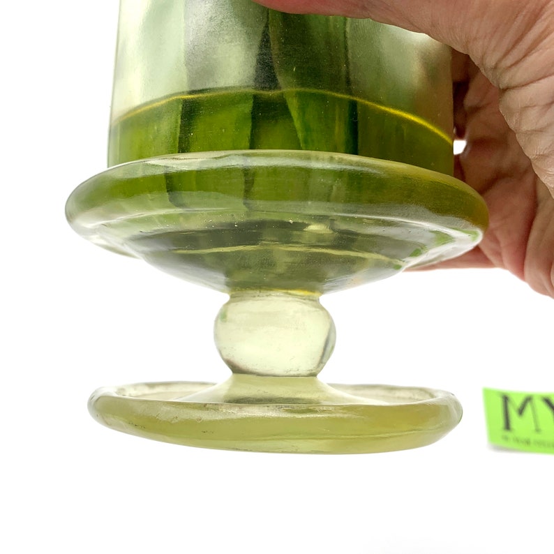 May include: A clear glass jar with a green tint and a wide base. The jar has a round, smooth knob on the bottom of the base.