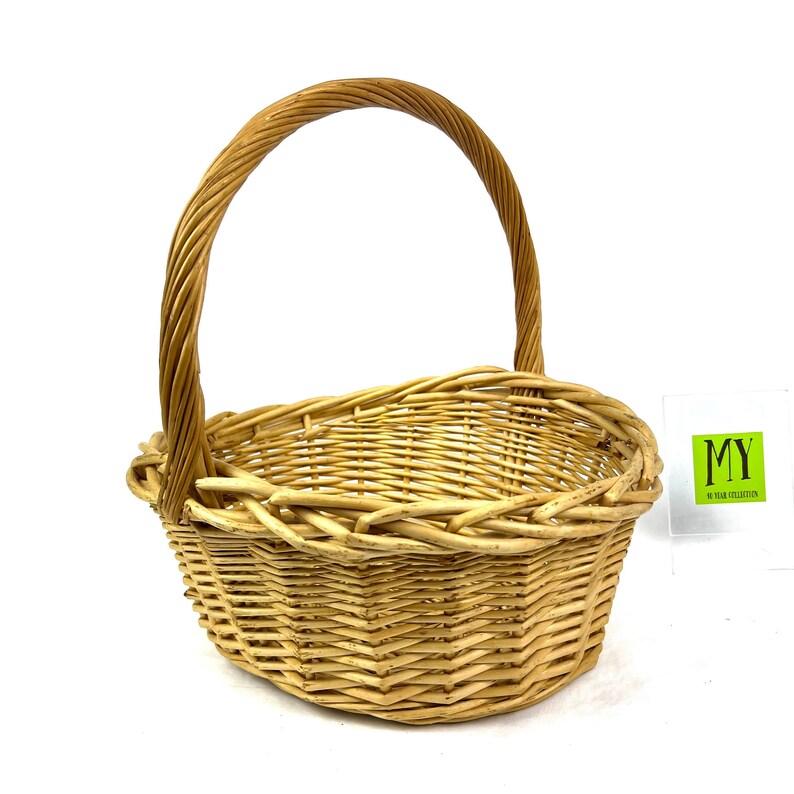 May include: A natural woven wicker basket with a rounded oval shape and a single handle. The basket is made of light brown wicker and has a natural, rustic look.