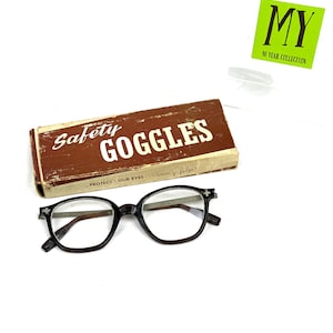 Pode incluir: &Oacute;culos de prote&ccedil;&atilde;o vintage com arma&ccedil;&atilde;o castanha escura e lentes transparentes, ao lado de uma caixa castanha com a inscri&ccedil;&atilde;o "Safety Goggles". A caixa tem um aspeto envelhecido. Tamb&eacute;m &eacute; vis&iacute;vel um autocolante verde com "MY 40 YEAR COLLECTION".