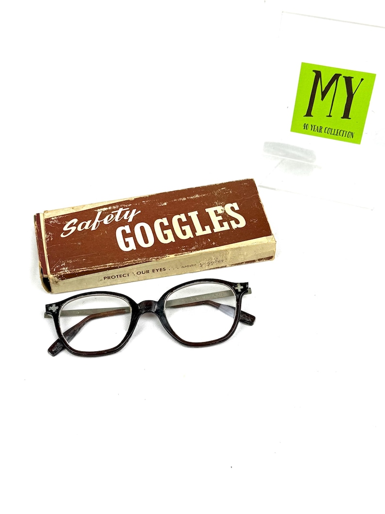 Pode incluir: &Oacute;culos de prote&ccedil;&atilde;o vintage com arma&ccedil;&atilde;o castanha escura e lentes transparentes, ao lado de uma caixa castanha com a inscri&ccedil;&atilde;o "Safety Goggles". A caixa tem um aspeto envelhecido. Tamb&eacute;m &eacute; vis&iacute;vel um autocolante verde com "MY 40 YEAR COLLECTION".