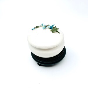 May include: A white ceramic trinket box with a floral design. The box has a round lid with a raised edge and is decorated with blue and green forget-me-not flowers. The box sits on a black wooden base.