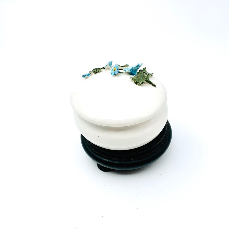 May include: A white ceramic trinket box with a floral design. The box has a round lid with a raised edge and is decorated with blue and green forget-me-not flowers. The box sits on a black wooden base.