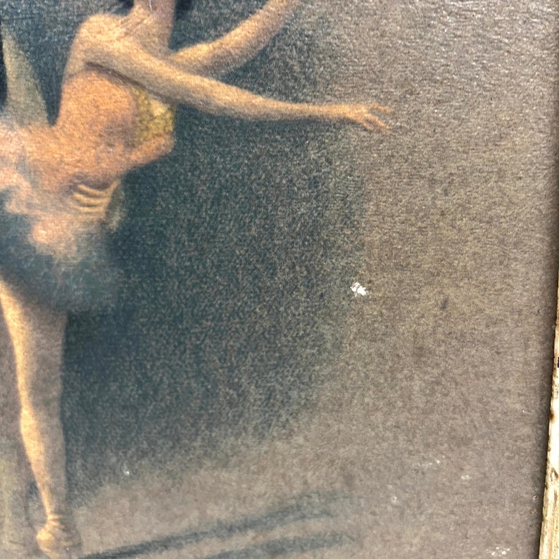 May include: A vintage framed print of a ballerina in a black tutu and a gold bodice, performing a dance pose. The background is a dark gray.