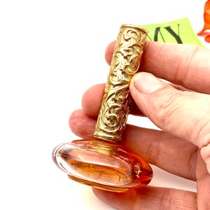 May include: A vintage perfume bottle with a gold, ornate, cylindrical stopper and a clear, amber-colored glass base.