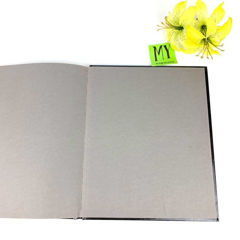 May include: A black hardcover book with a gray interior, open to a blank page. A green sticky note with the text "MY 50 YEAR COLLECTION" is visible on the top right corner of the book.