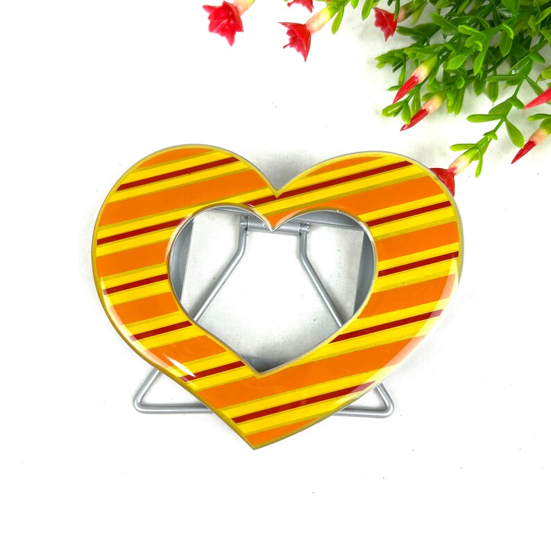 May include: A heart-shaped metal stand with a yellow and orange striped design. The stand is designed to hold a picture or other object.