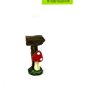 May include: A miniature wooden sign with an arrow pointing to the right, reading "Welcome". A red and white mushroom with white spots is attached to the sign post.