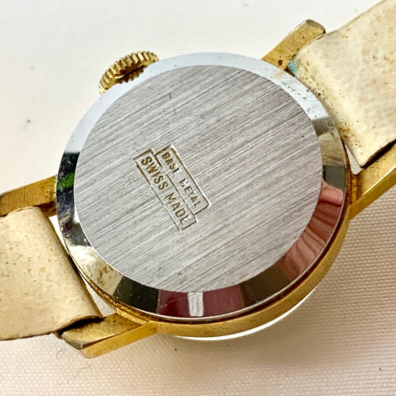 May include: Close-up of a vintage wristwatch, showcasing the back of the watch. The watch has a gold-toned metal case and a cream-colored leather strap. The watch back is silver with the text "BASE METAL" and "SWISS MADE".