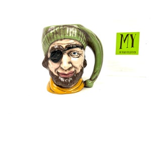 May include: A green and brown ceramic mug shaped like a pirate's head with an eye patch. The mug has a green hat and a yellow scarf. The mug is part of the "MY 40 YEAR COLLECTION" series.
