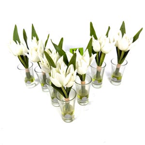Peut inclure: Plusieurs bouquets de tulipes blanches dans des verres &agrave; shot transparents. Chaque verre contient plusieurs tulipes avec des feuilles vertes, attach&eacute;es avec de la ficelle. Les verres &agrave; shot sont dispos&eacute;s en forme de V sur fond blanc.