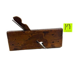 Könnte beinhalten: Ein alter hölzerner Handhobel mit einer Metallklinge. Der Hobel besteht aus dunkelbraunem Holz und hat einen rechteckigen Körper mit einem gebogenen Griff. Die Klinge ist abgewinkelt und ragt von der Seite des Hobels heraus. Das Bild enthält den Text "MY" in einem grünen Feld.