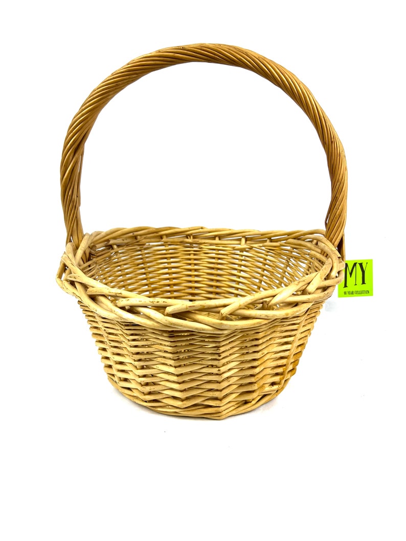 May include: A natural woven wicker basket with a single handle. The basket is light brown in color and has a rounded shape.