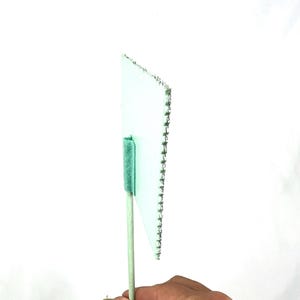 May include: A small, light green flag with a white border and a row of silver staples along one edge. The flag is attached to a light green wooden stick with a green fuzzy material at the top. The flag is held by a hand.