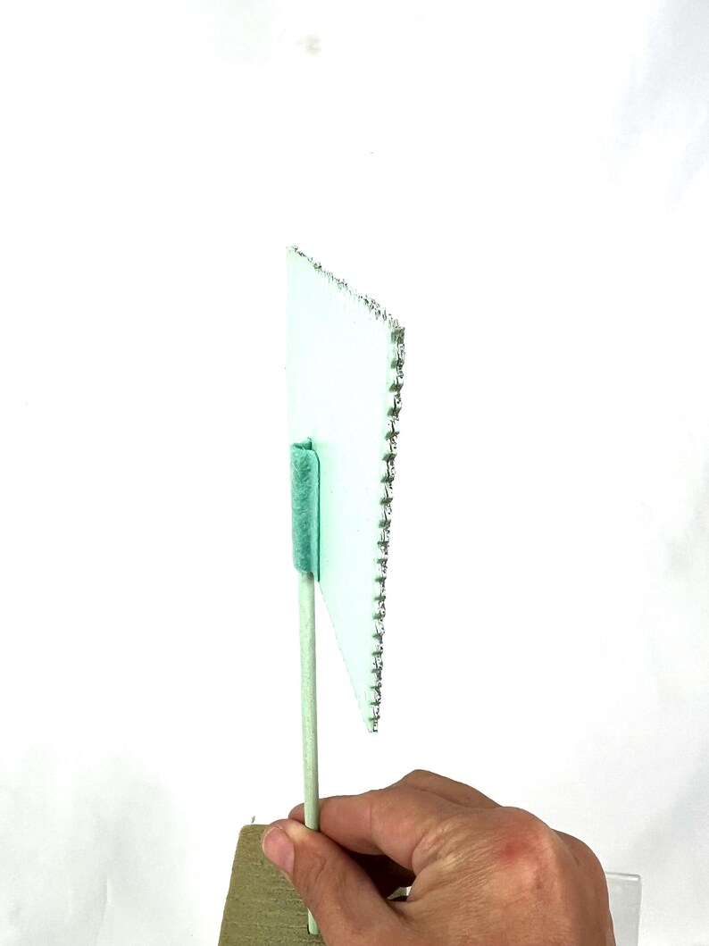 May include: A small, light green flag with a white border and a row of silver staples along one edge. The flag is attached to a light green wooden stick with a green fuzzy material at the top. The flag is held by a hand.