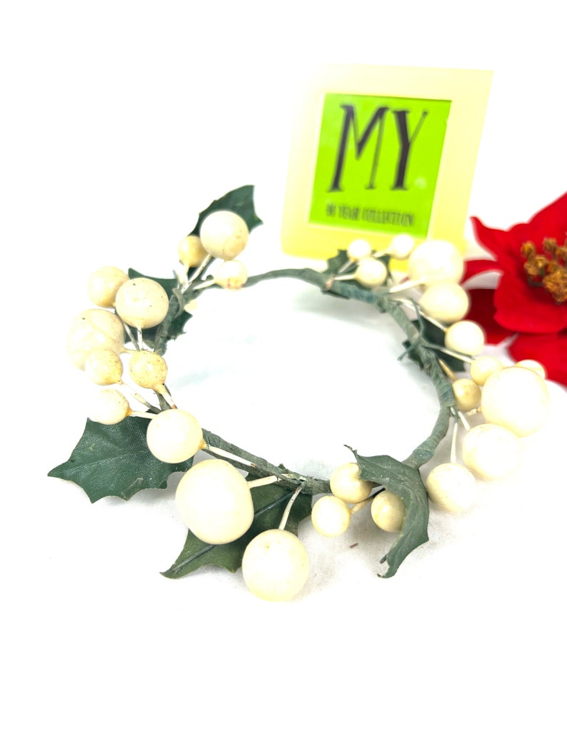 May include: A white and green artificial wreath with white berries and green leaves. The wreath is made of wire and is about 10 inches in diameter. The wreath is sitting on a white surface. The text "MY YEAR COLLECTION" is visible on a yellow tag.