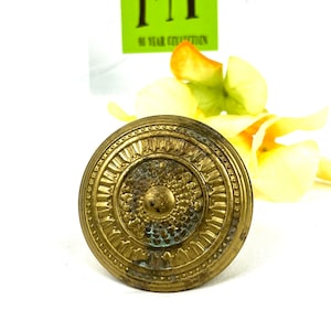 Könnte beinhalten: Ein runder, goldfarbener Metallknopf mit einem erhabenen, verzierten Design. Der Knopf hat eine kreisförmige Mitte mit einer strukturierten Oberfläche und einen Ring aus erhabenen, spitzen Blütenblättern darum herum.