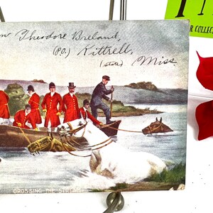 K&ouml;nnte beinhalten: Eine alte Postkarte, die eine Gruppe von M&auml;nnern in roten M&auml;nteln zeigt, die auf Pferden einen Bach &uuml;berqueren. Die Postkarte tr&auml;gt den Titel "Crossing the Stream" und ist an Theodore Breland in Kittrell, Mississippi, adressiert.