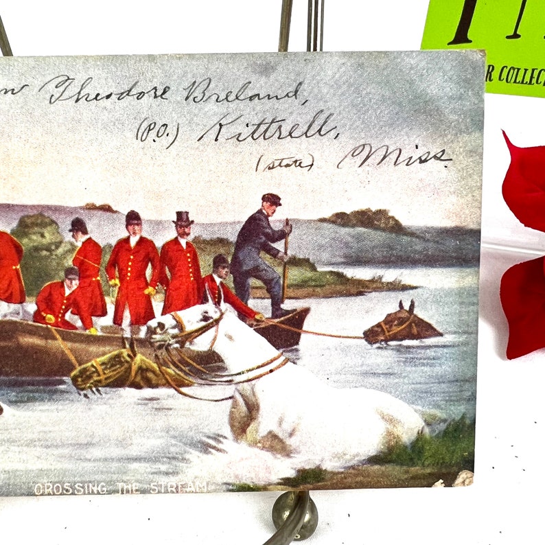 K&ouml;nnte beinhalten: Eine alte Postkarte, die eine Gruppe von M&auml;nnern in roten M&auml;nteln zeigt, die auf Pferden einen Bach &uuml;berqueren. Die Postkarte tr&auml;gt den Titel "Crossing the Stream" und ist an Theodore Breland in Kittrell, Mississippi, adressiert.
