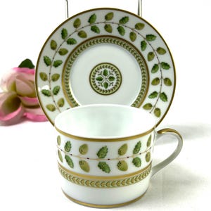 May include: A white porcelain teacup and saucer with a green and gold floral pattern. The cup has a handle and the saucer has a scalloped edge.