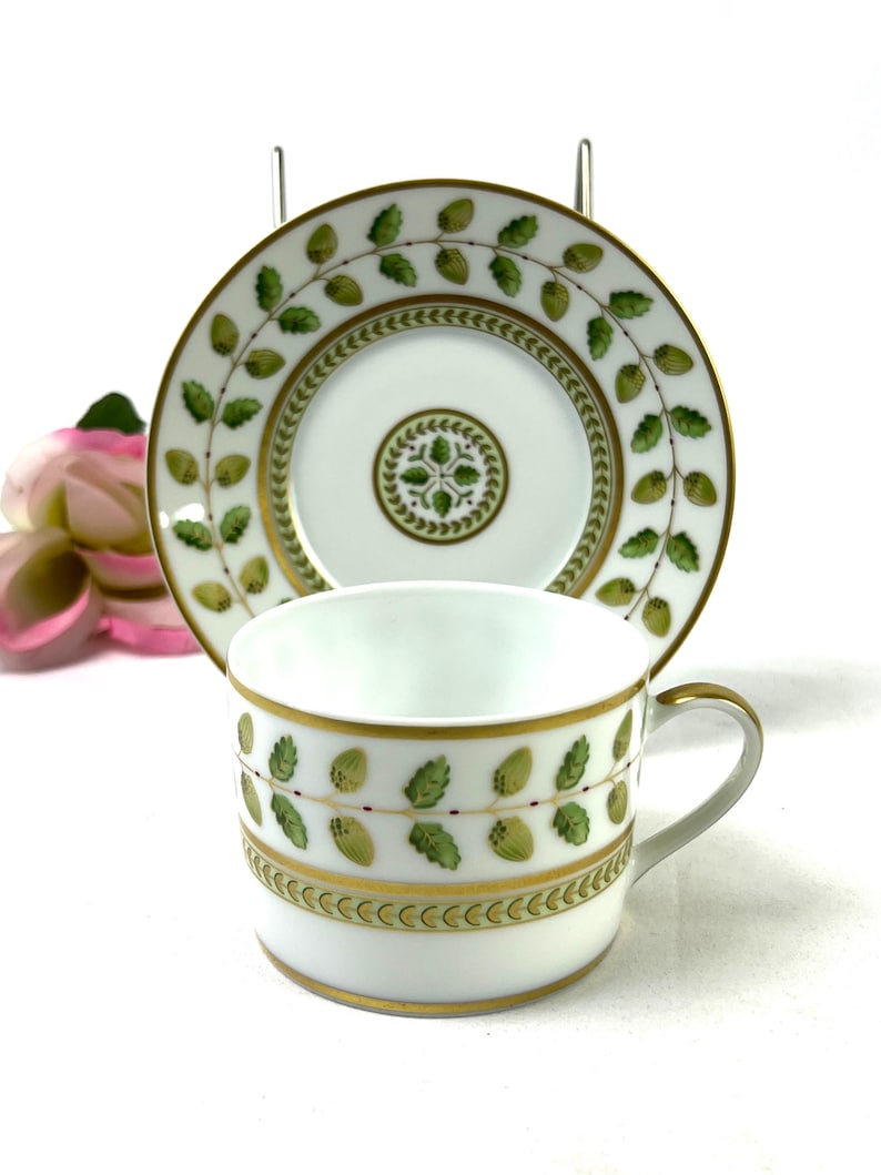 May include: A white porcelain teacup and saucer with a green and gold floral pattern. The cup has a handle and the saucer has a scalloped edge.