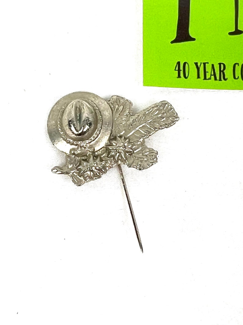 May include: Silver-toned pin featuring a hat with a feather and leaves. The pin is attached to a long, thin pin.