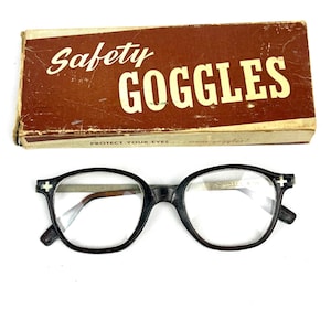 Pode incluir: &Oacute;culos de prote&ccedil;&atilde;o pretos vintage com lentes transparentes, em frente a uma caixa de papel&atilde;o marrom. A caixa diz "Safety Goggles" em escrita branca. Os &oacute;culos t&ecirc;m um design de arma&ccedil;&atilde;o redondo cl&aacute;ssico.