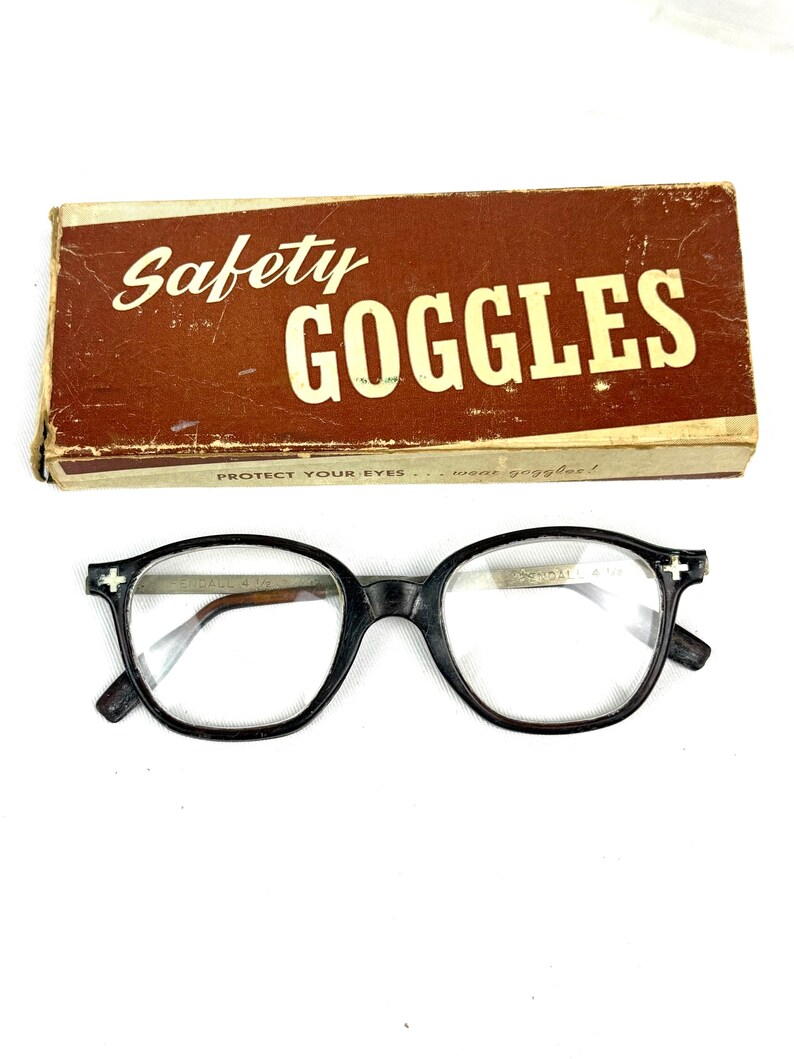 Pode incluir: &Oacute;culos de prote&ccedil;&atilde;o pretos vintage com lentes transparentes, em frente a uma caixa de papel&atilde;o marrom. A caixa diz "Safety Goggles" em escrita branca. Os &oacute;culos t&ecirc;m um design de arma&ccedil;&atilde;o redondo cl&aacute;ssico.