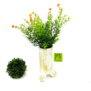 May include: A white ceramic planter with three legs and a green leafy plant with orange flowers. The planter has a distressed white finish and the text "MY HOME COLLECTION" is on a white tag attached to the planter.