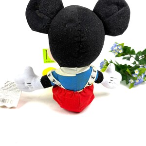 May include: A Mickey Mouse plush toy, viewed from the back, with black ears, white gloves, and red shorts. The toy has a blue shirt with white suspenders and the letter "W" on it. The toy is on a white background.