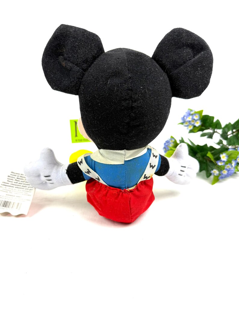 May include: A Mickey Mouse plush toy, viewed from the back, with black ears, white gloves, and red shorts. The toy has a blue shirt with white suspenders and the letter "W" on it. The toy is on a white background.