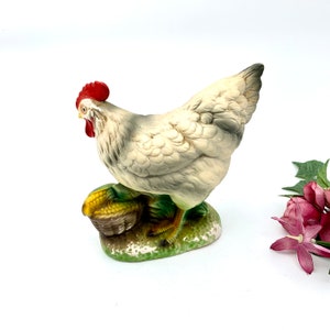 May include: White ceramic figurine of a hen with a red comb, standing on a green base with a basket of corn.