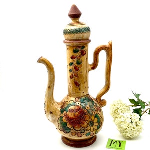 May include: A ceramic teapot with a floral design in shades of brown, green, and orange. The teapot has a tall, slender body and a lid with a pointed top. The teapot is decorated with a floral pattern that includes roses, leaves, and other flowers.