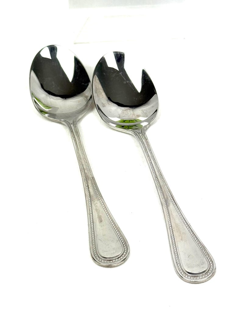 May include: Two shiny, silver serving spoons with a slotted design. The spoons have a teardrop-shaped bowl and a long handle with a beaded border. The spoons are likely for serving salad or other dishes.