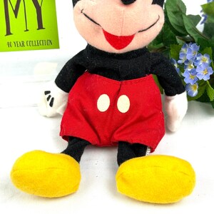 May include: A plush Mickey Mouse doll with a black body, red shorts with white buttons, and yellow shoes. The doll has a smiling face with a red mouth and white gloves. A green tag with the text "MY 40 YEAR COLLECTION" is visible.