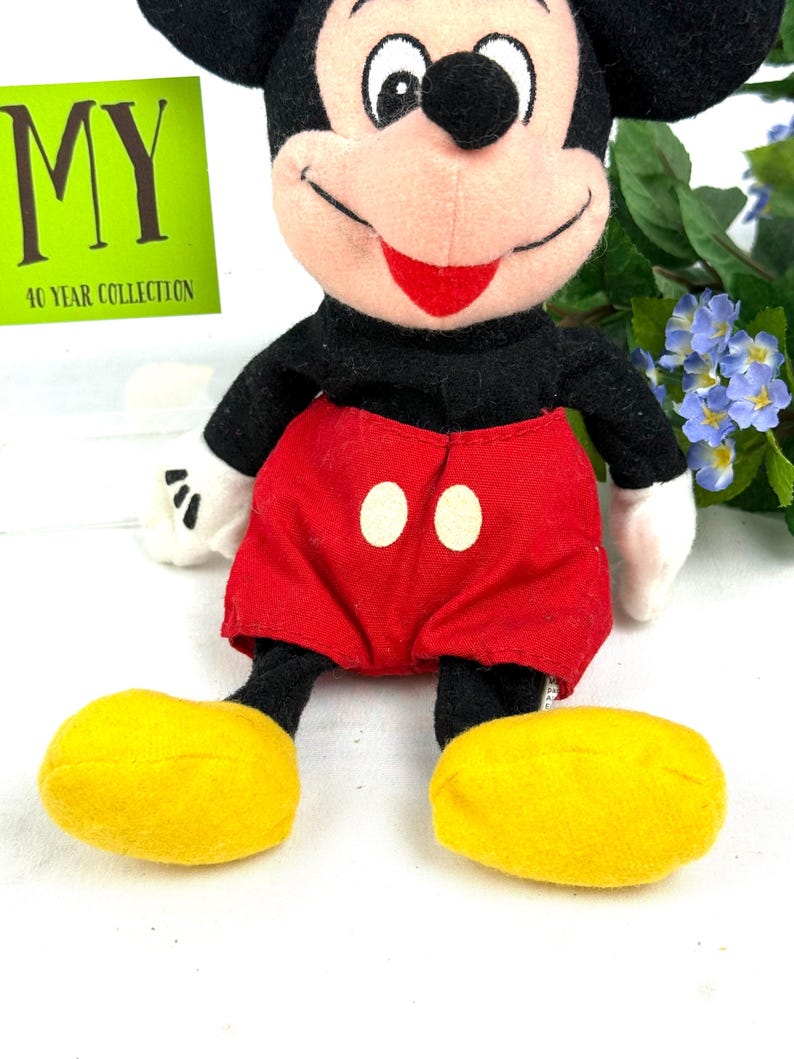 May include: A plush Mickey Mouse doll with a black body, red shorts with white buttons, and yellow shoes. The doll has a smiling face with a red mouth and white gloves. A green tag with the text "MY 40 YEAR COLLECTION" is visible.