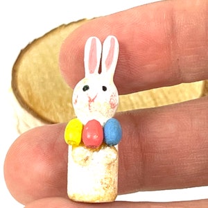 May include: A white wooden bunny figurine with pink ears and a yellow, pink, and blue Easter egg garland around its neck.