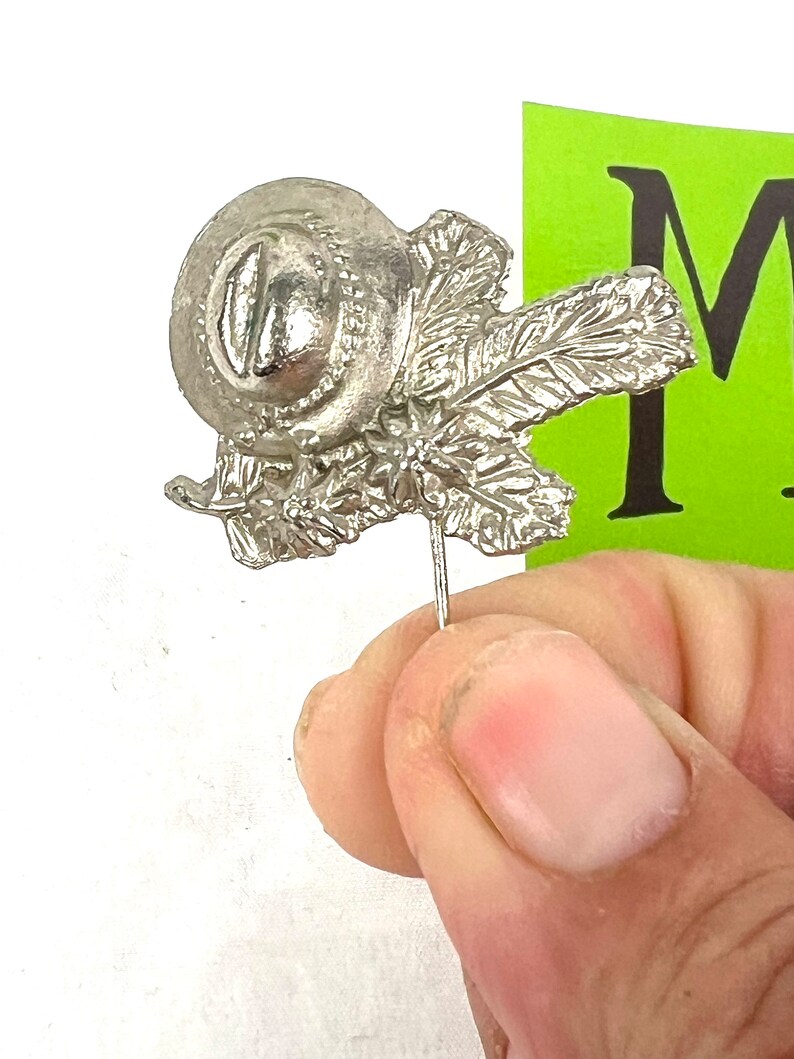 May include: A silver pin with a detailed design of a round fruit with leaves. The pin is attached to a thin metal rod.