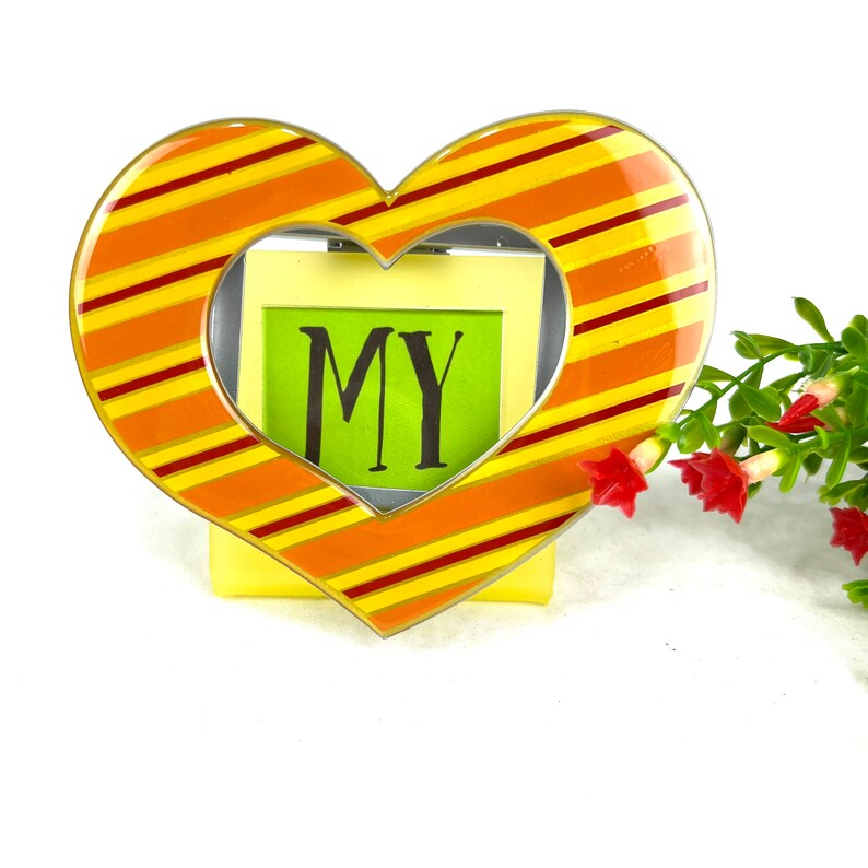 May include: A heart-shaped picture frame with yellow and orange stripes. The frame has a clear opening for a photo and the word "MY" is written on a green paper inside the frame.