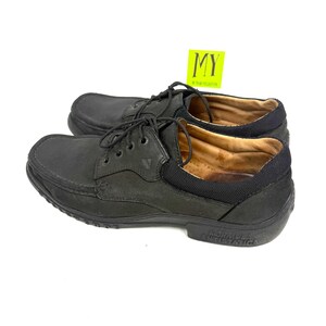 May include: A pair of black leather boat shoes with laces. The shoes have a rounded toe and a black rubber sole. The text "FORMULA ANTISTATICA" is visible on the sole. A yellow tag with the letters "MY" is in the background.