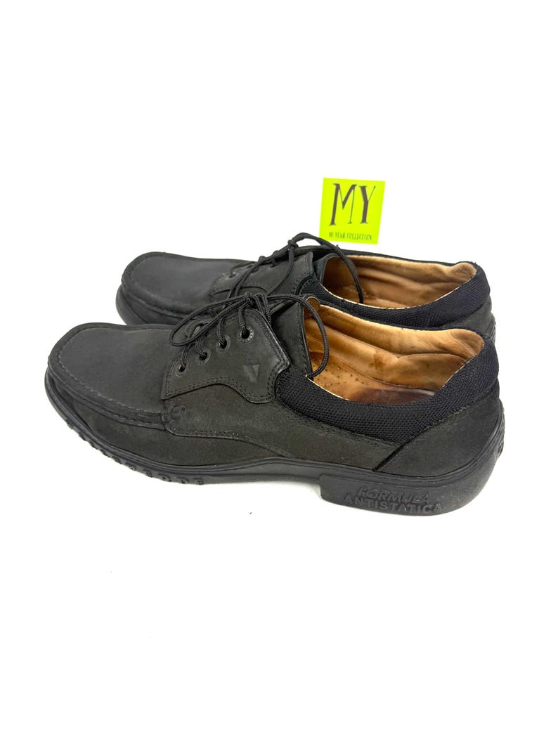 May include: A pair of black leather boat shoes with laces. The shoes have a rounded toe and a black rubber sole. The text "FORMULA ANTISTATICA" is visible on the sole. A yellow tag with the letters "MY" is in the background.