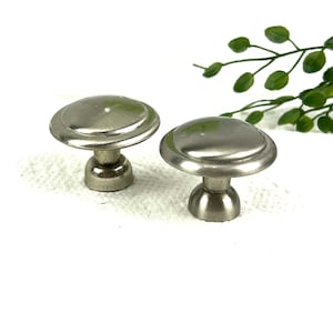 May include: Two silver cabinet knobs with a round, slightly raised top and a round base. The knobs are on a white surface.