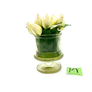 May include: A clear glass vase with a pedestal base holds a bouquet of white tulips and green leaves. The vase has a textured surface. The vase is labeled "MY 50 YEAR COLLECTION".