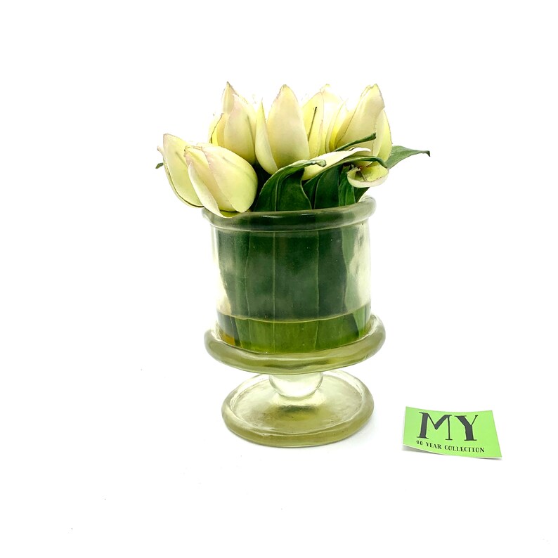May include: A clear glass vase with a pedestal base holds a bouquet of white tulips and green leaves. The vase has a textured surface. The vase is labeled "MY 50 YEAR COLLECTION".
